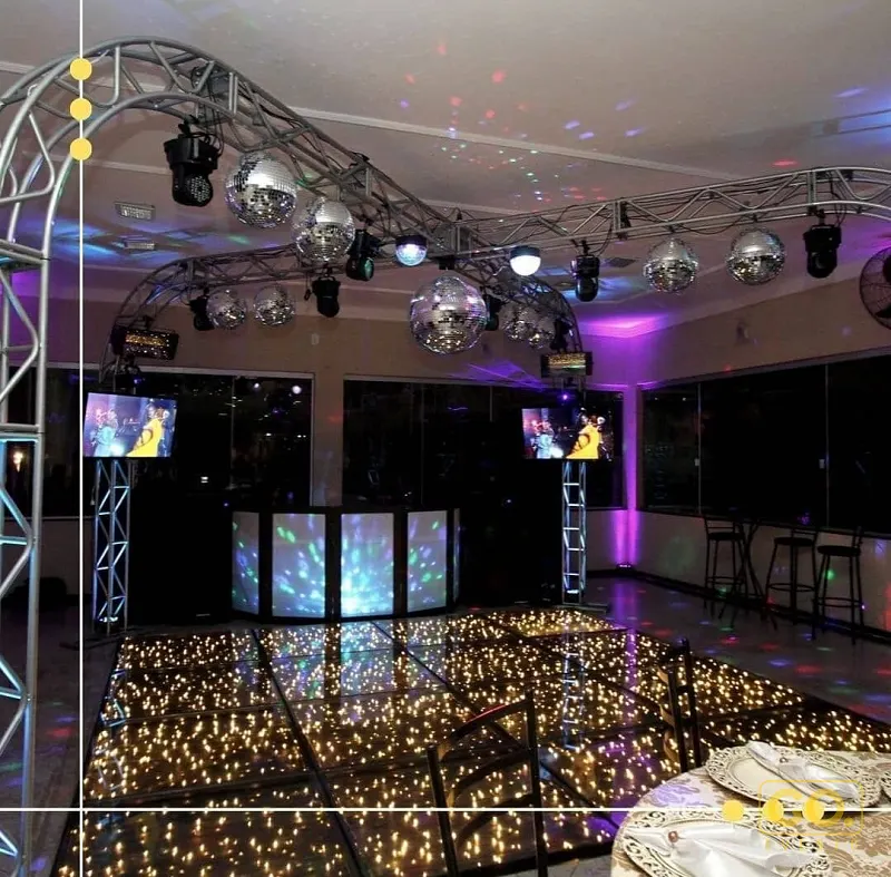 Salão de festas à noite vazio, com bolas de espelho, treliça iluminada, cabine de DJ com painéis de LED e pista de luzes.