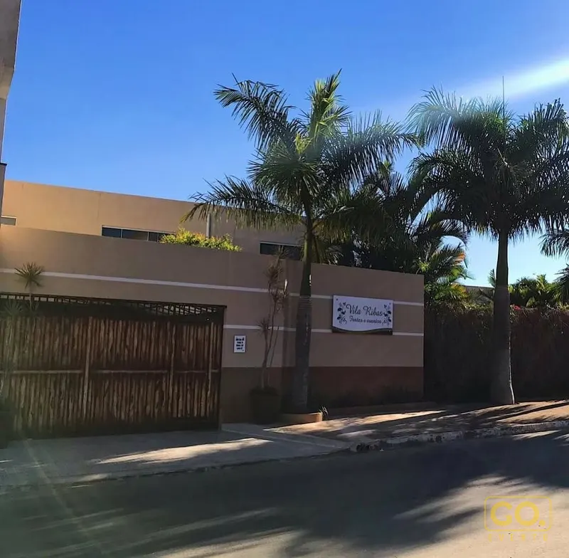 Fachada de muro bege com portão de bambu e palmeiras altas sob céu azul, em rua arborizada durante o dia.