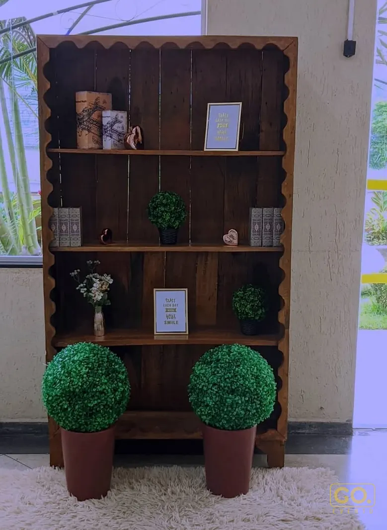 Estante de madeira com bordas onduladas decorada com plantas artificiais, porta-retratos e livros sobre tapete branco.