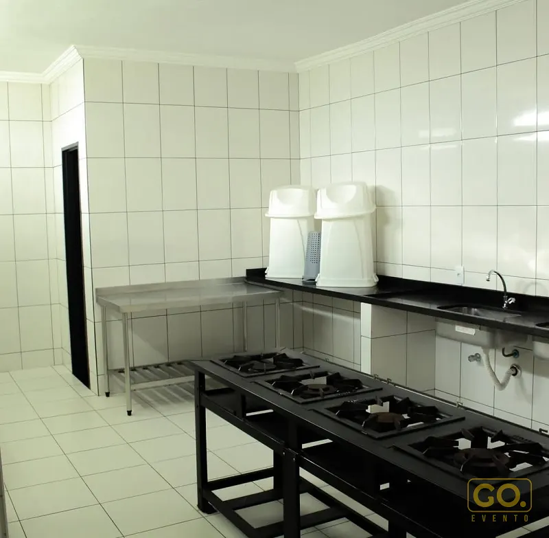 Cozinha industrial com fogão de quatro bocas preto, bancada de inox e azulejos brancos, sem pessoas à vista.