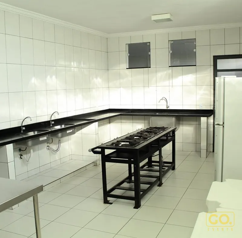 Cozinha industrial vazia com fogão de quatro bocas em metal preto, bancada de granito e azulejos brancos nas paredes.