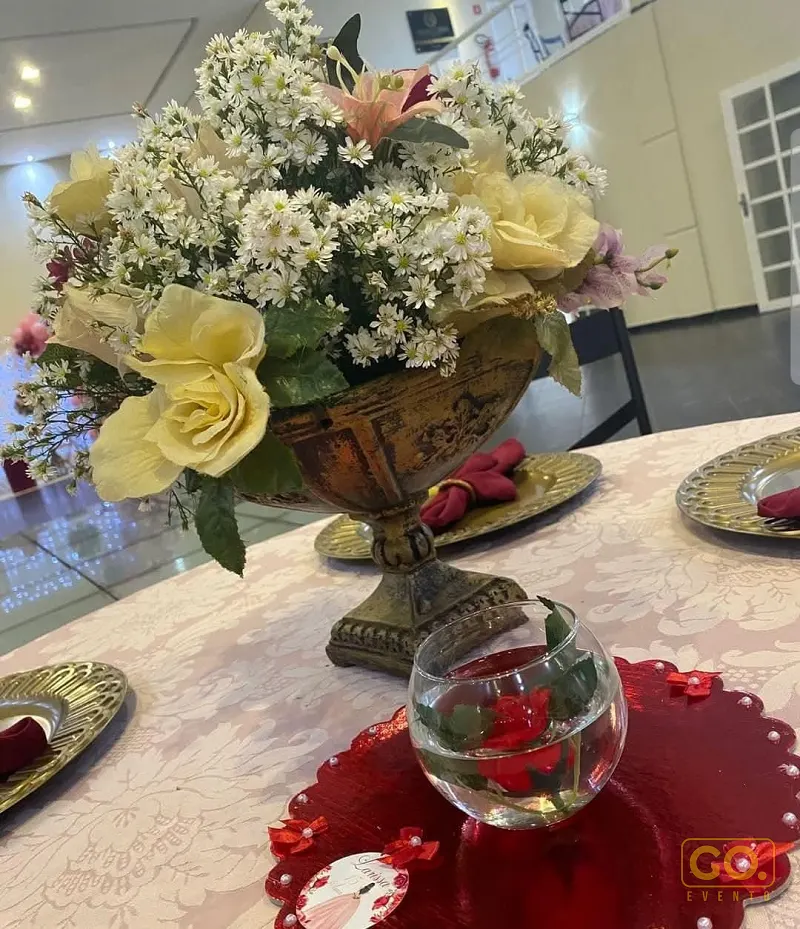 Mesa posta com arranjo de flores brancas e amarelas em urna dourada, pratos dourados e sousplat vermelho com pérolas.