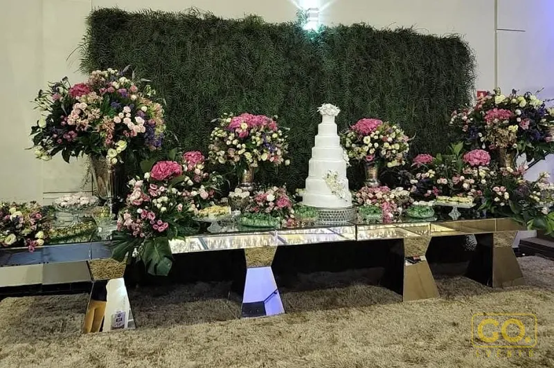 Mesa de doces espelhada com bolo branco de vários andares, arranjos de flores rosas e roxas e parede verde ao fundo.