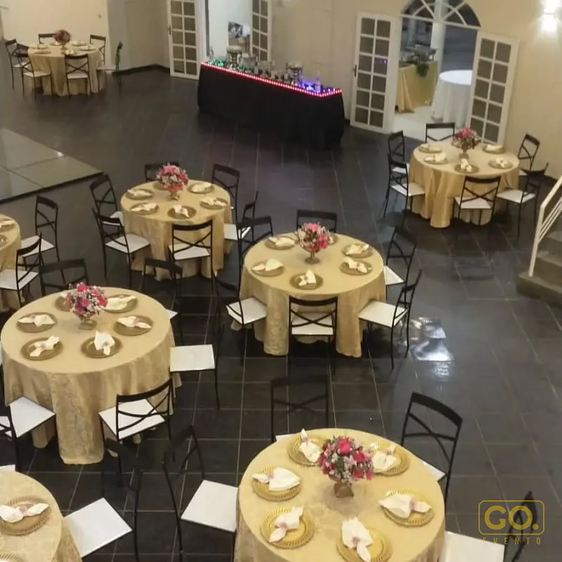 Salão de festas com mesas redondas de toalha dourada, cadeiras pretas e arranjos de flores rosa, montado para evento sem convidados.