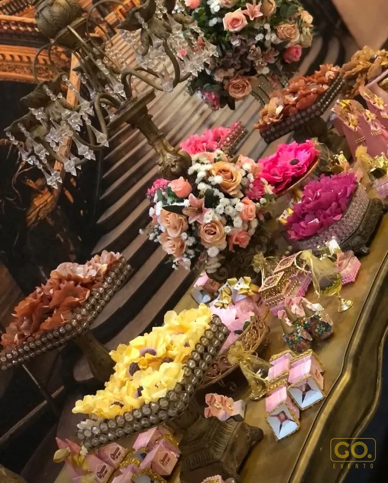 Mesa de doces decorada com arranjos de flores rosa, amarelo e pêssego, suportes dourados com strass e lembrancinhas.