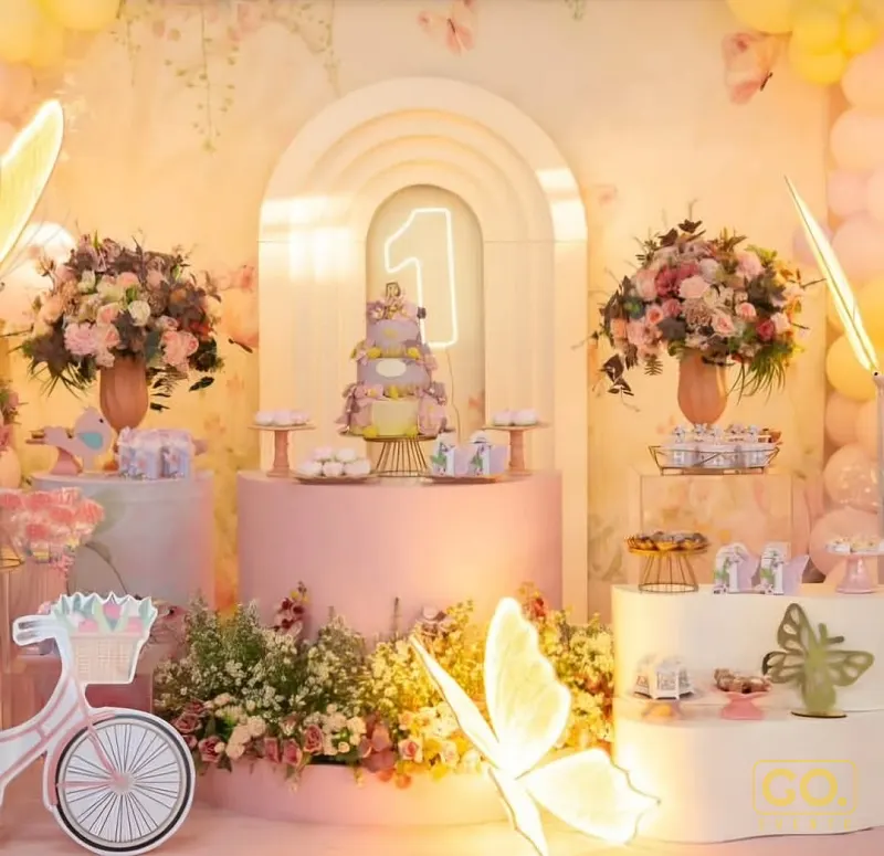 Painel de festa infantil em tons pastel com arco, bolo de dois andares, flores, balões e um grande número 1 em neon.