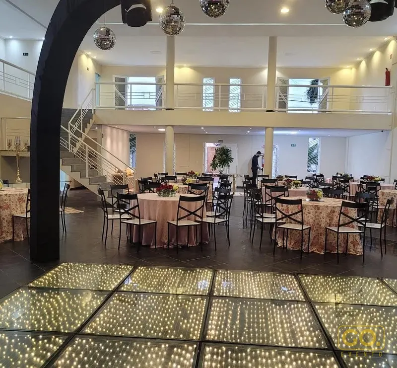 Salão de festas com mesas redondas vestidas em toalhas rosê, cadeiras pretas e piso de LED iluminado em primeiro plano.