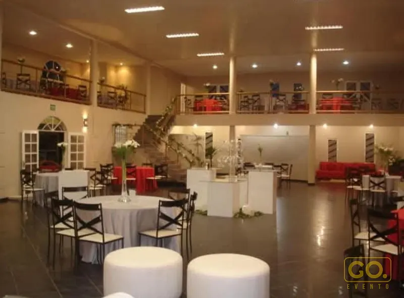 Salão de eventos com mesas redondas de toalha branca, mezanino com mesas ao redor e escada ao fundo, sem convidados.
