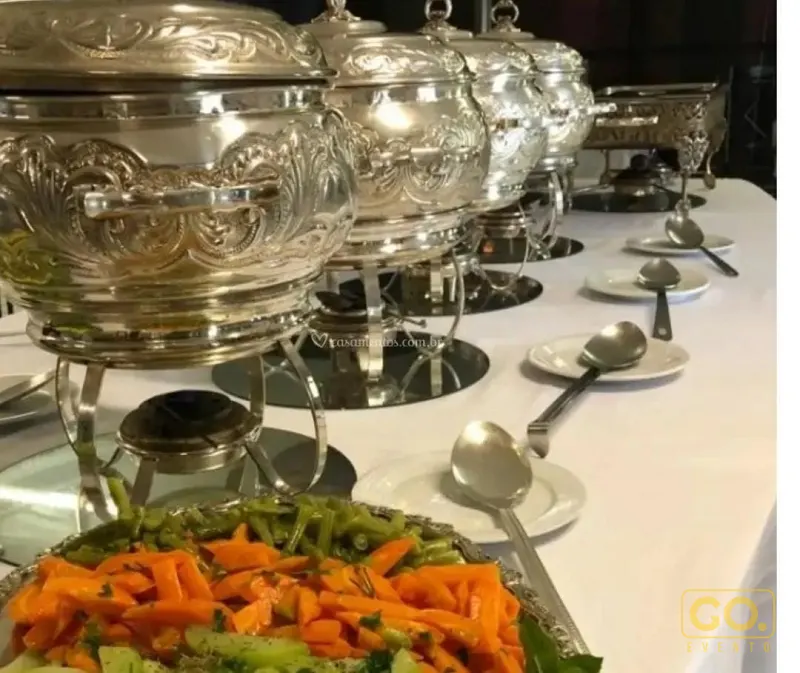 Mesa de buffet com cubas térmicas de prata cinzeladas, pratos brancos, colheres de servir e legumes refogados em primeiro plano.