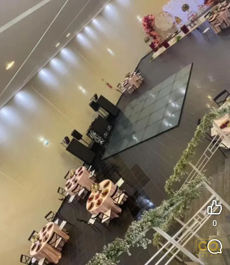 Salão de eventos montado com mesas redondas de toalha rosa, pista espelhada e guirlandas verdes ao longo da escada, visto do alto.