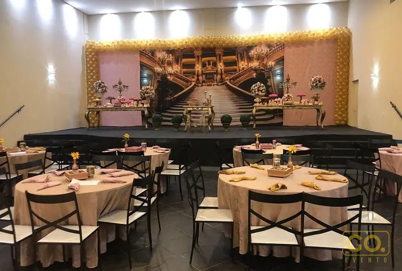 Salão decorado com mesas redondas de toalhas bege, palco com painel de cenário de ópera e arco de balões dourados ao fundo.