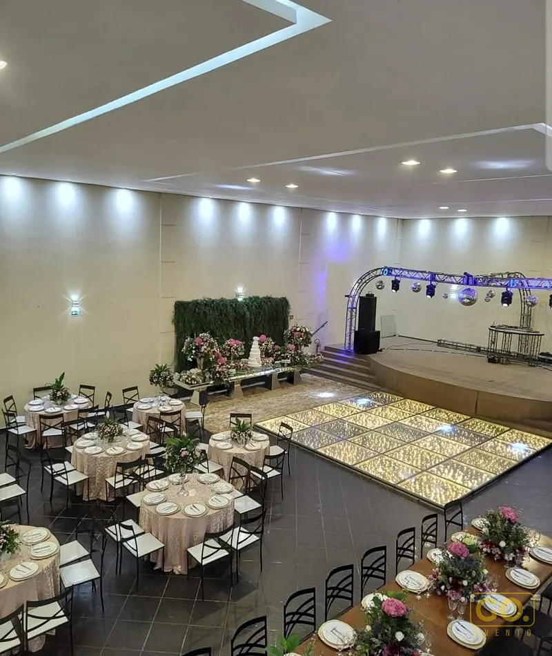 Salão de festas com mesas redondas vestidas, mesa de bolo decorada com flores, palco com treliça e pista de dança iluminada.