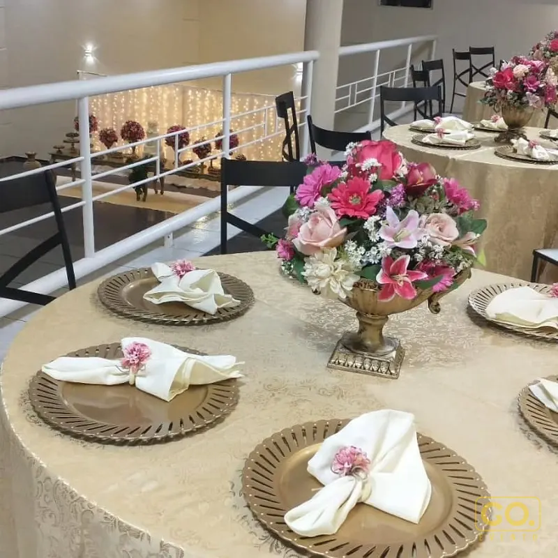 Salão montado com mesas redondas de toalha bege, pratos dourados, arranjo de flores cor-de-rosa e luzes de fada ao fundo.