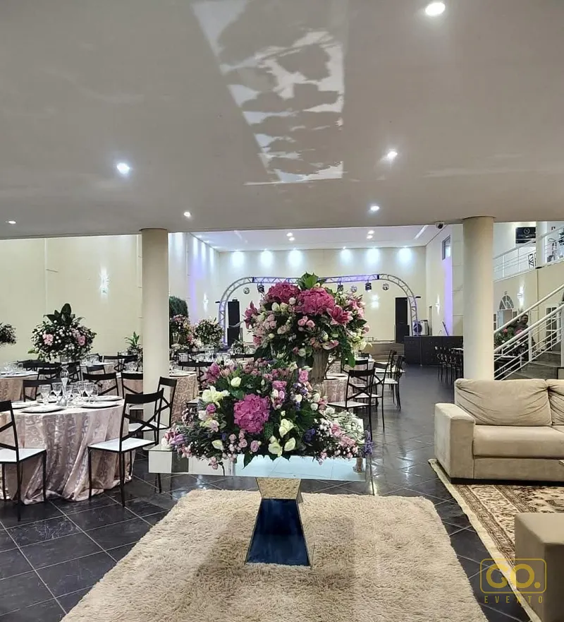 Salão de festas coberto com mesas redondas postas com toalhas rosé, arranjos de flores e iluminação de spots no teto.