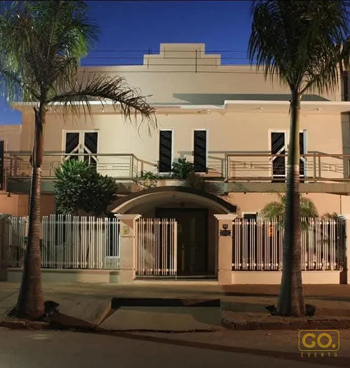 Fachada de sobrado de dois andares à noite, com sacadas de grade metálica, portão de madeira sob arco e palmeiras na calçada.
