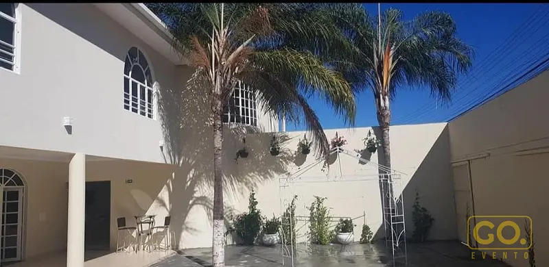 Pátio externo de piso cinza com duas palmeiras, treliças de ferro com plantas e uma mesa com cadeiras sob área coberta.