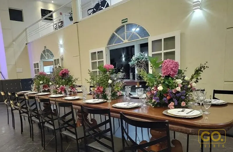 Salão com mesa comprida de madeira posta com pratos, taças e arranjos de flores rosas e brancas, sob pé-direito alto.