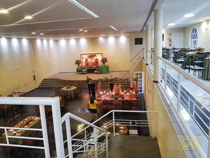 Vista elevada de salão de festas com palco ao fundo, mesas redondas com toalhas verdes e mesa decorada com velas e flores.