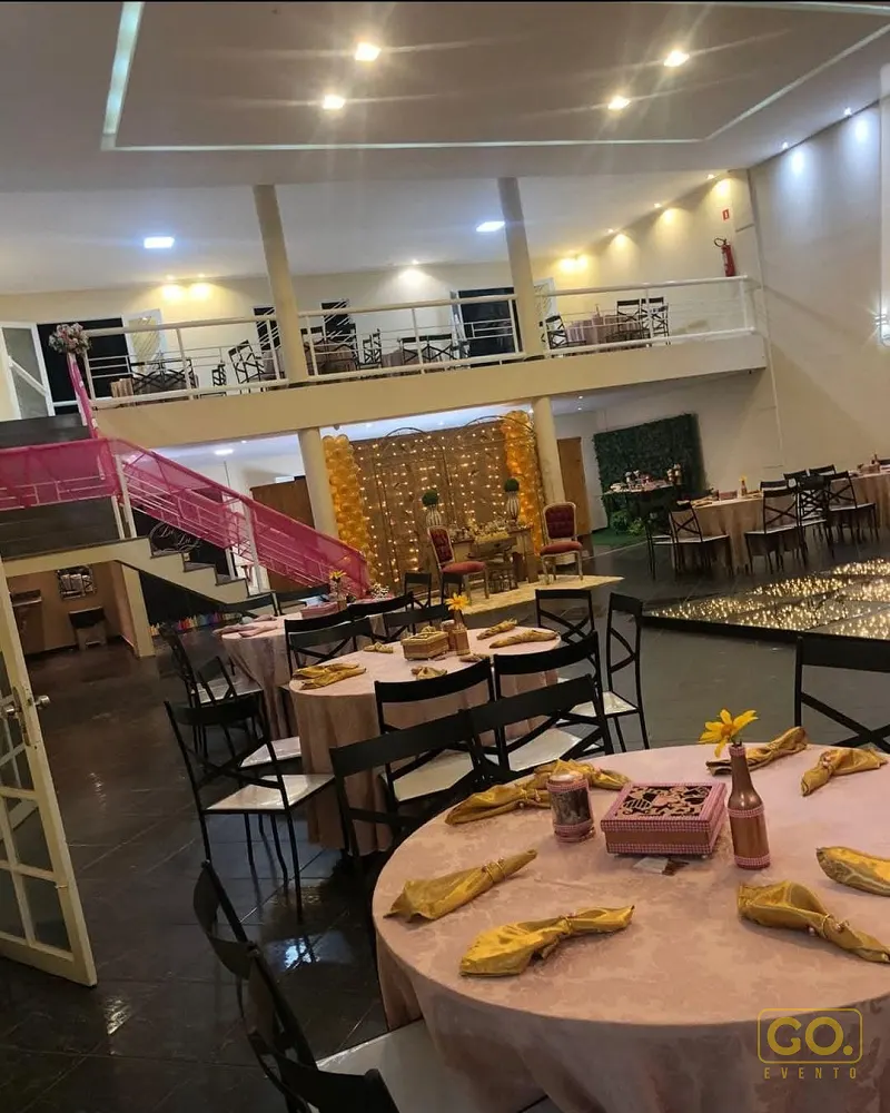 Salão de festas com mesas redondas postas com toalhas rosa, guardanapos amarelos e mezanino ao fundo, sem convidados.