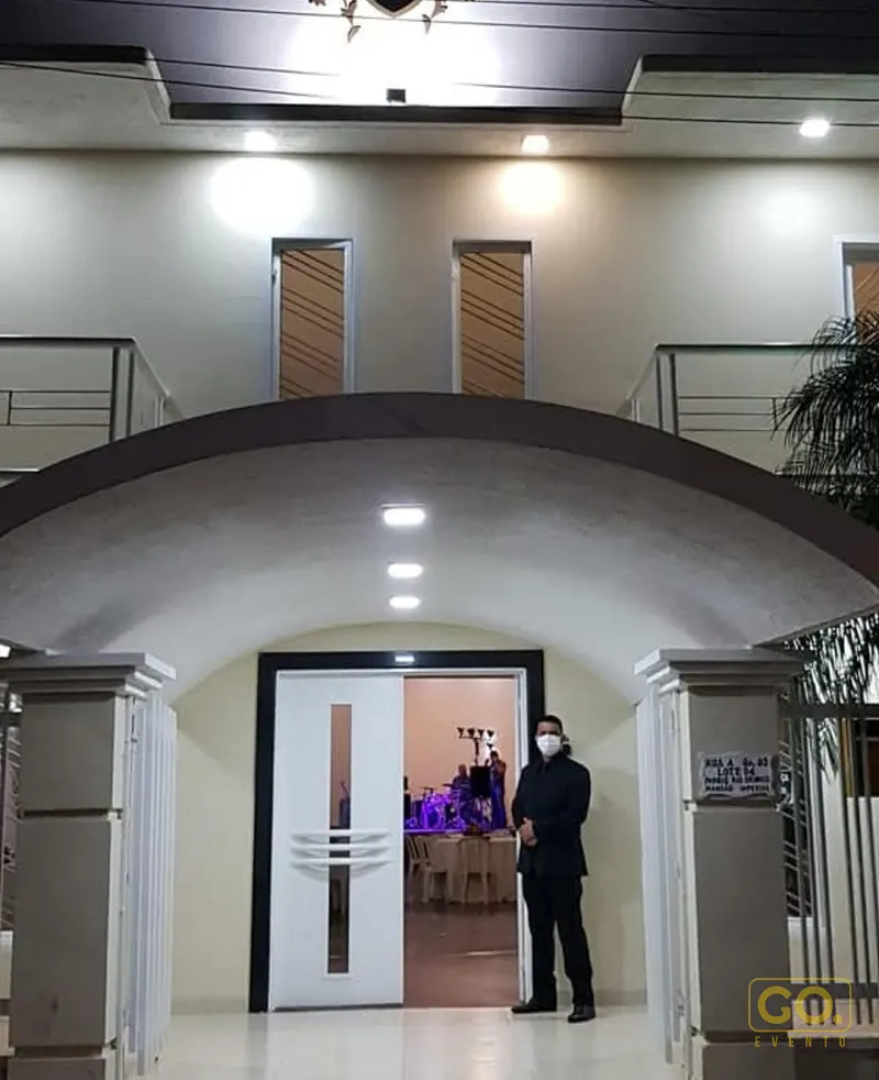 Fachada de sobrado com entrada em arco iluminada à noite, com porta aberta revelando mesas postas e bateria ao fundo do salão.