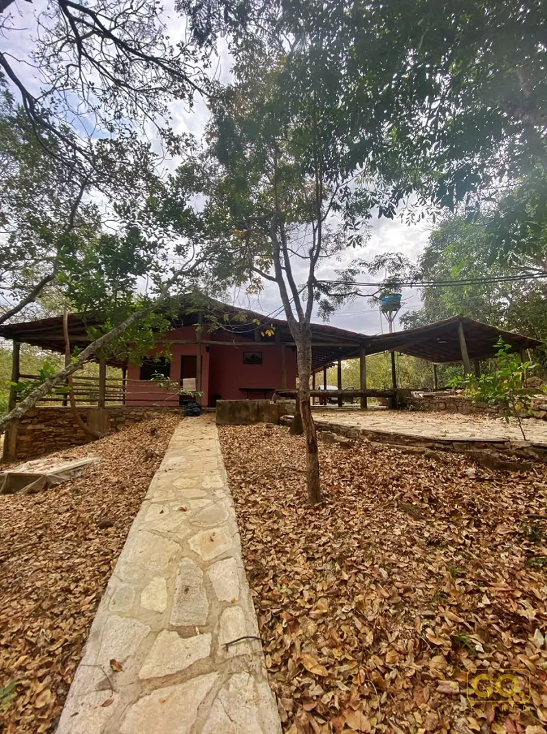 Casa rústica de paredes avermelhadas com varanda coberta por pérgola de madeira, cercada de árvores e caminho de pedra.
