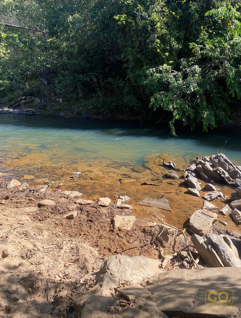 Margem de rio com água azulada e transparente, pedras e terra exposta em primeiro plano, cercada por mata densa e árvores.