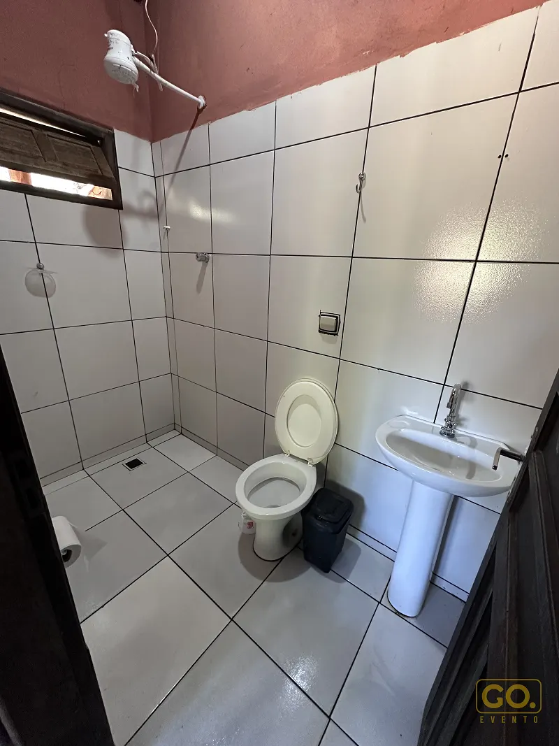 Banheiro simples com azulejos brancos, vaso sanitário, pia de coluna e chuveiro elétrico aparente, sem nenhum evento montado.