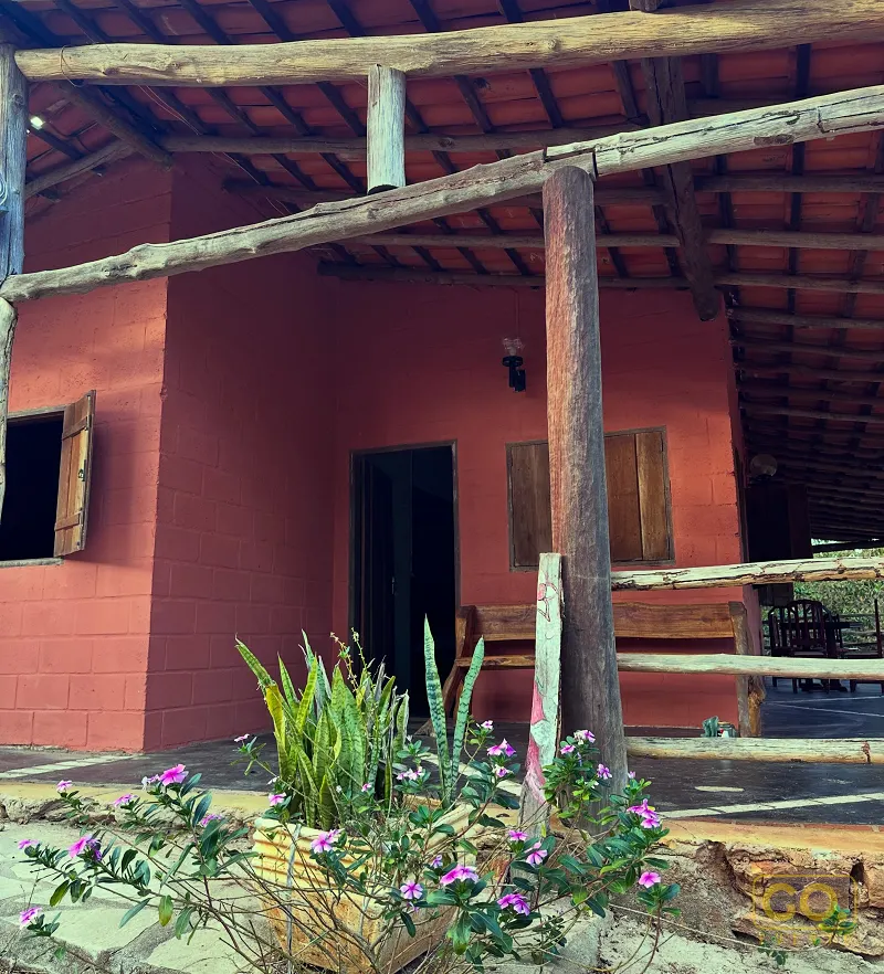 Varanda coberta com pilares e vigas de madeira, paredes vermelhas e vasos de plantas com flores cor-de-rosa na entrada.