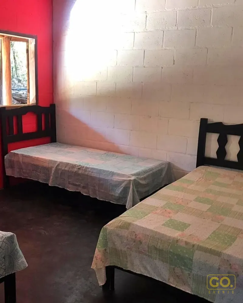 Quarto simples com duas camas de solteiro, colchas de retalho, parede vermelha e janela de madeira com vista para a mata.