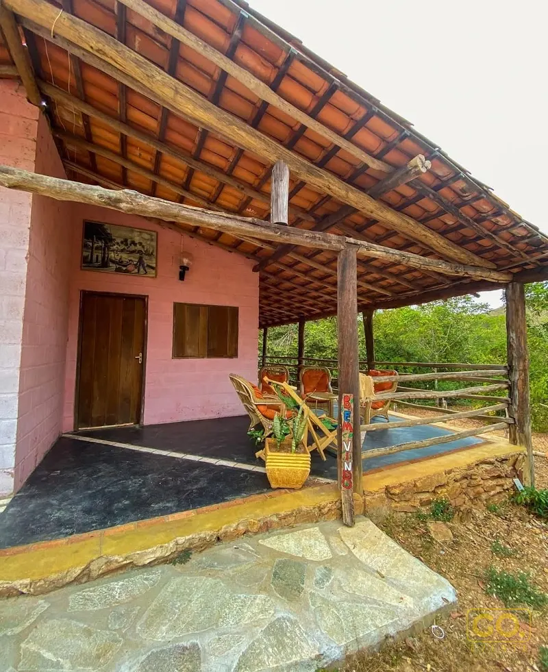 Varanda coberta rústica com cadeiras de vime, vaso de planta e parede rosa, com vista para vegetação verde ao fundo.