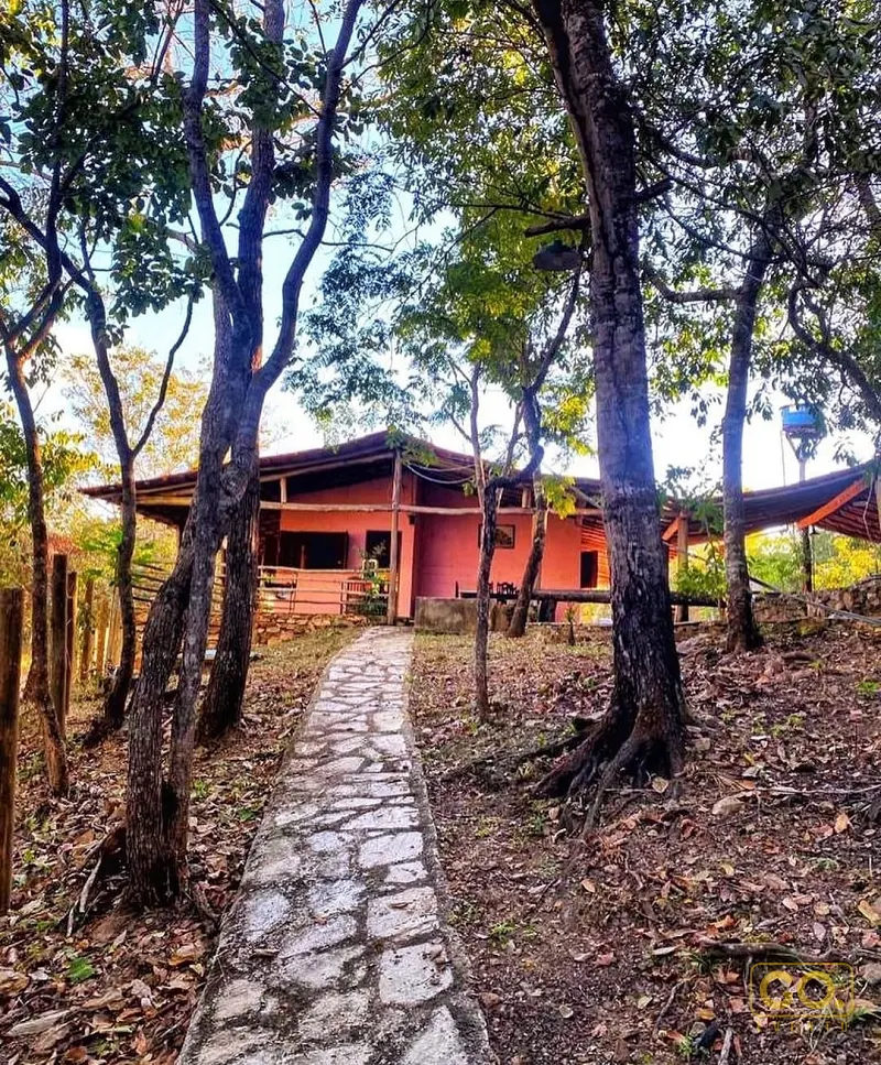 Casa rústica de cor coral em meio a árvores, com caminho de pedra irregular levando até a entrada e varanda de madeira.
