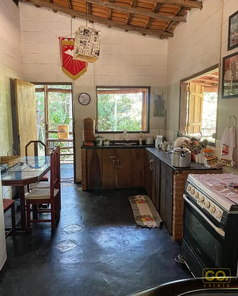Cozinha rústica com paredes de bloco, fogão preto, pia com bancada escura e mesa de vidro com cadeiras ao fundo, vista para a mata.