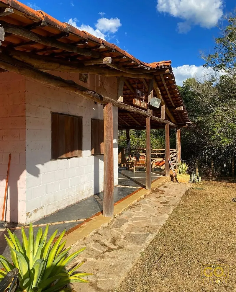 Varanda coberta de uma casa rústica com telhado de barro, pilares de madeira e caminho de pedra cercado por mata.