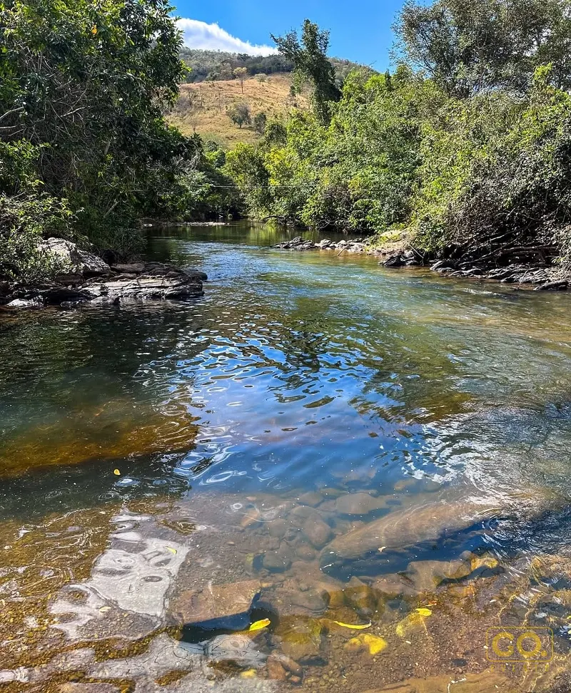 Rio de águas claras e pedras no leito, cercado por mata verde e um morro de vegetação seca sob céu azul.