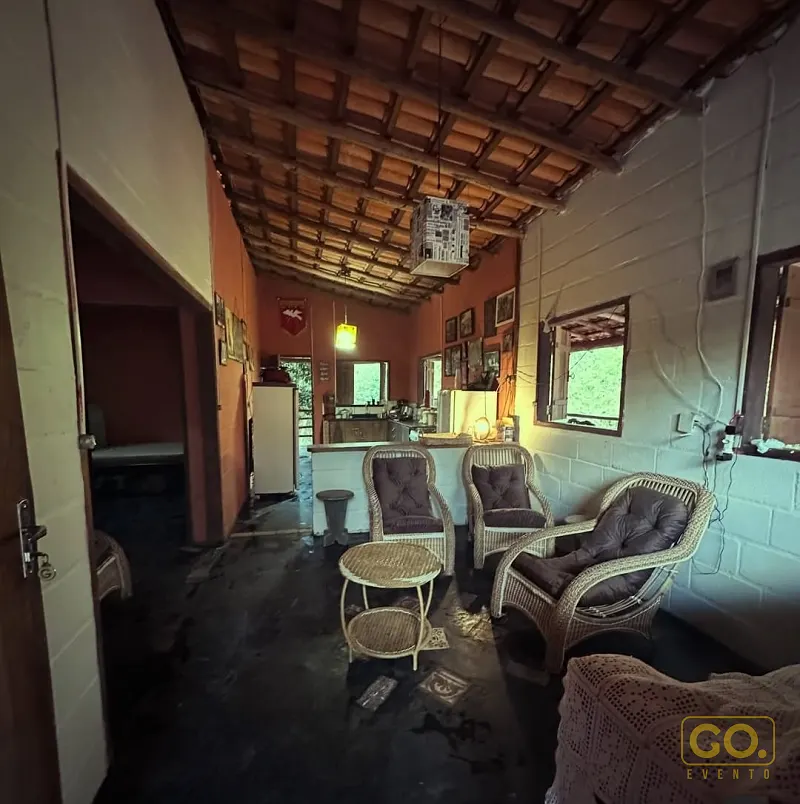 Sala rústica com poltronas de vime almofadadas, mesa de centro de vime, teto de vigas de madeira e cozinha ao fundo, sem pessoas.