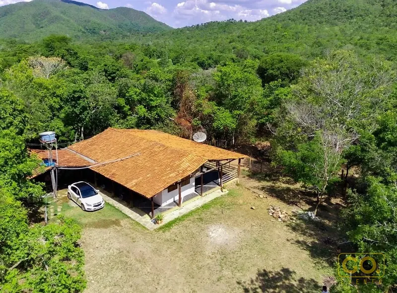 Vista aérea de casa rústica com telhado de telha cerâmica, varanda e terreno cercado por mata e serras verdes.