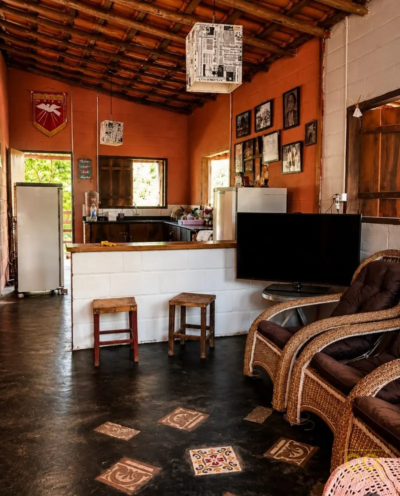 Sala rústica com paredes laranjas, poltronas de vime, cozinha ao fundo e piso de cimento queimado com ladrilhos decorativos.