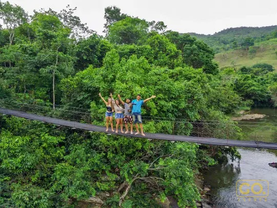 Vista aérea de ponte suspensa sobre rio em meio à mata, com um grupo de quatro pessoas de braços erguidos.