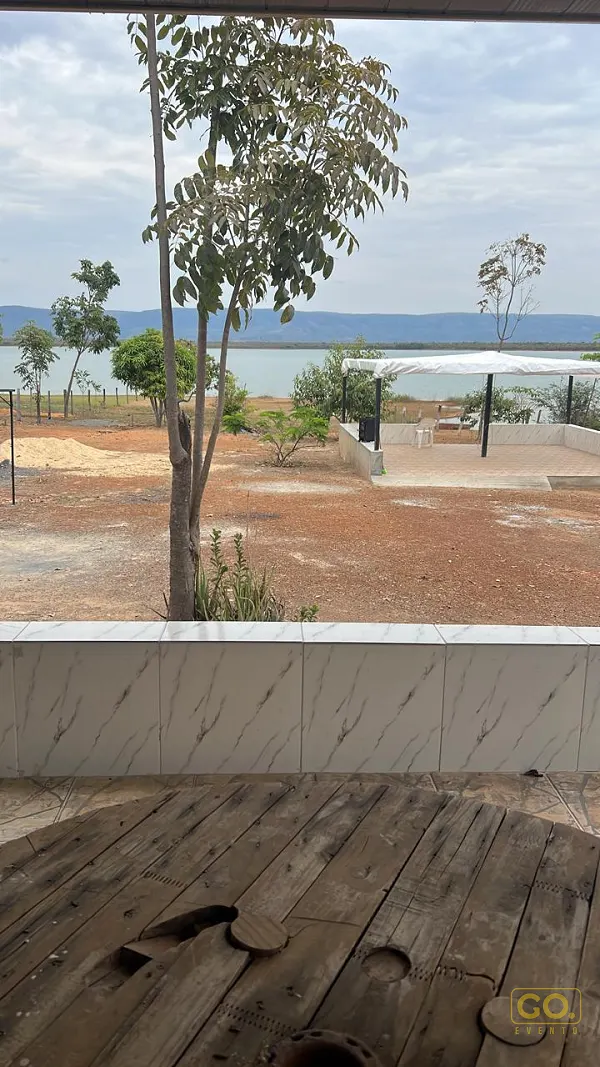 Vista de área externa com piso de terra e pérgola branca voltada para um lago com montanhas ao fundo, sob céu nublado.