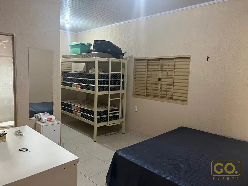 Quarto simples com beliche de metal, cama de casal com colcha escura, mesa branca e janela com veneziana de madeira.