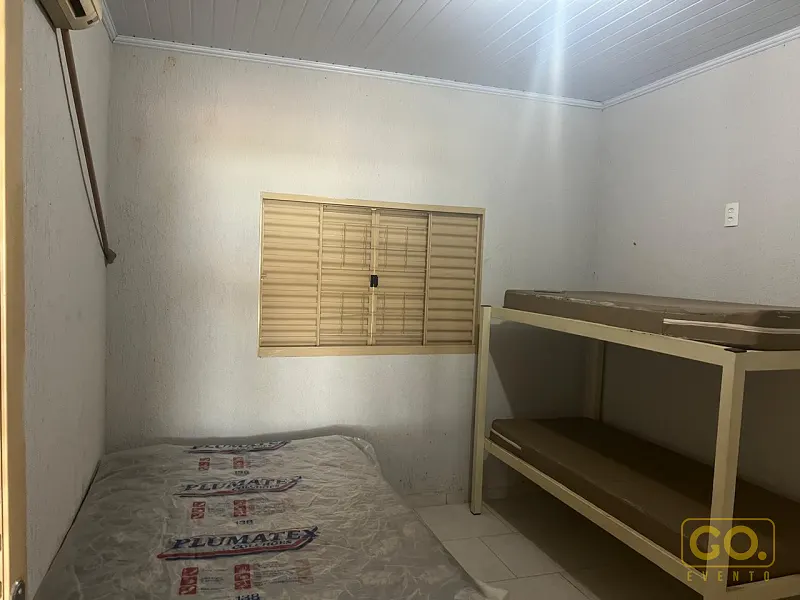 Quarto simples com cama de solteiro, beliche de metal e janela com veneziana, sem pessoas nem decoração de evento visível.