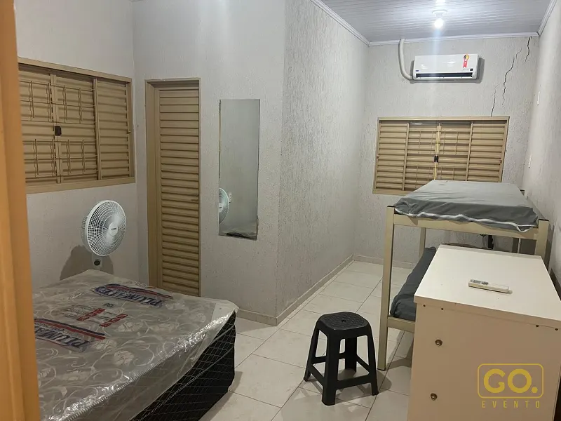 Quarto simples com cama de solteiro encapada, beliche, cômoda branca, ventilador e ar condicionado na parede.