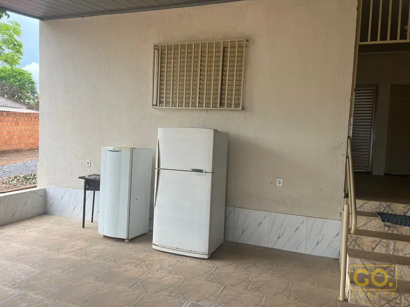 Área coberta com piso de cerâmica bege, duas geladeiras brancas, uma pequena churrasqueira e escada com corrimão ao lado da casa.