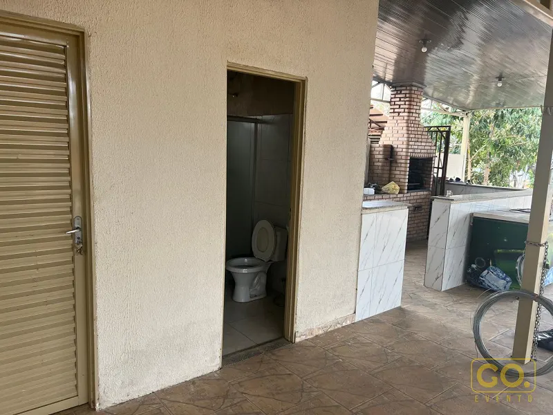 Área coberta com churrasqueira de tijolos aparentes e bancada de mármore, ao lado de uma porta aberta para banheiro com vaso sanitário.