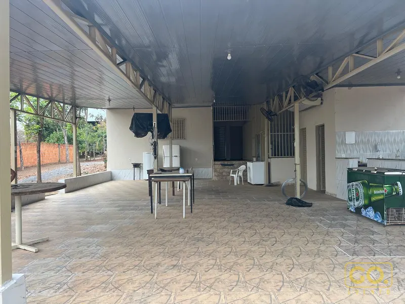 Área coberta com piso de cerâmica, mesas, cooler de bebidas e balcão com pia, aberta para o quintal e as árvores ao fundo.