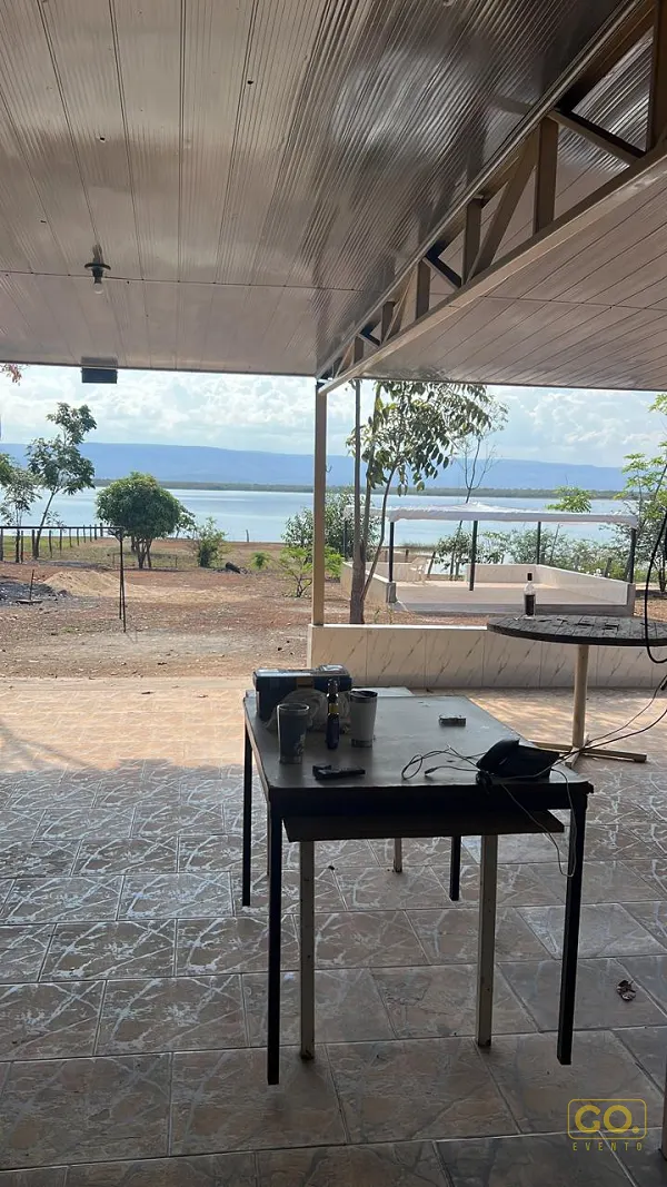Varanda coberta com piso de cerâmica e mesas, com vista para o lago e a serra ao fundo em dia de céu parcialmente nublado.