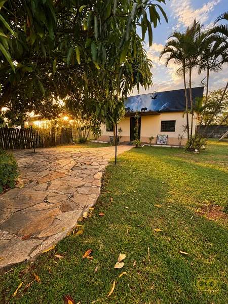Caminho de pedra irregular atravessa gramado até uma casa de paredes claras com telhado escuro, sob palmeiras ao entardecer.