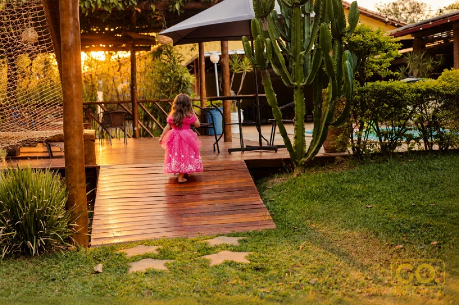 Menina de costas caminha por deck de madeira que leva a um gramado com cactos e piscina ao fundo.