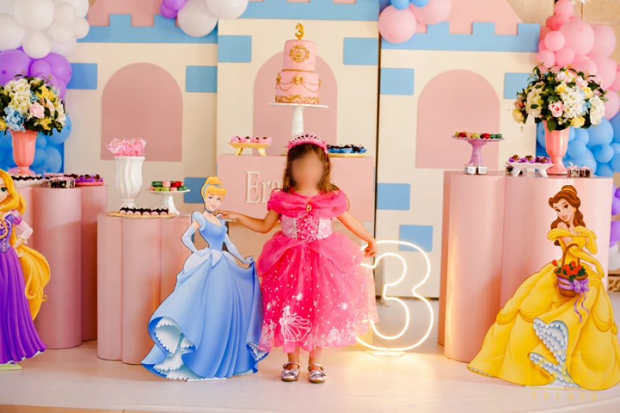 Criança de vestido rosa posa entre personagens de princesas de papel, bolo decorado e número 3 em neon numa festa infantil.