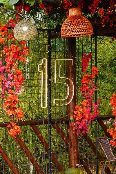 Detalhe de decoração ao ar livre com número 15 em neon, flores pendentes em vermelho e laranja e luminárias de palha entre a vegetação.